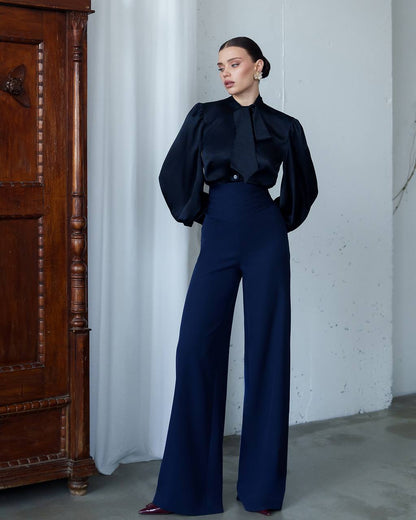 Woman in a black top and blue pants standing against a white wall with a wooden cabinet.