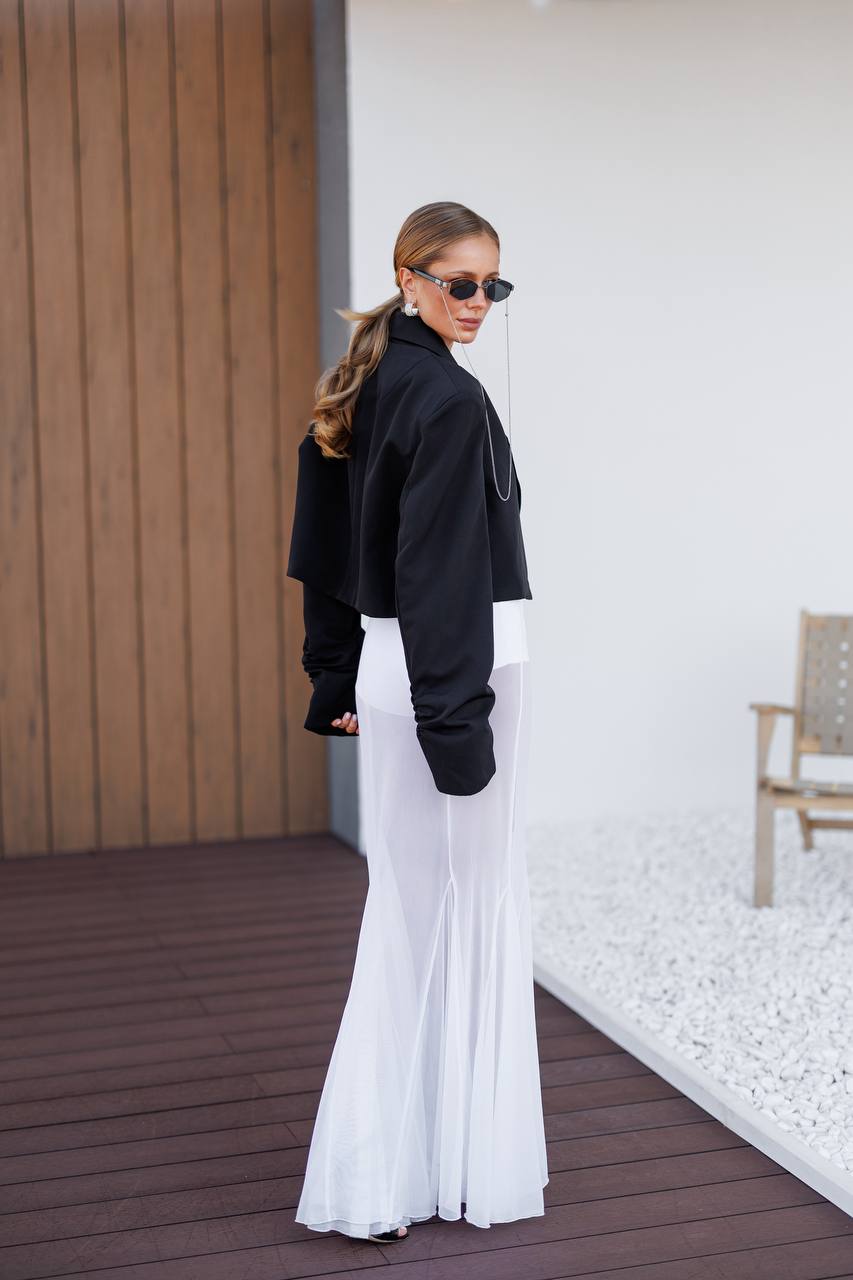 A woman stands on a wooden deck wearing a black cropped jacket, black gloves, and a long white skirt. She is photographed from the back in a modern outdoor setting with a white wall, gravel, and a wooden chair visible.