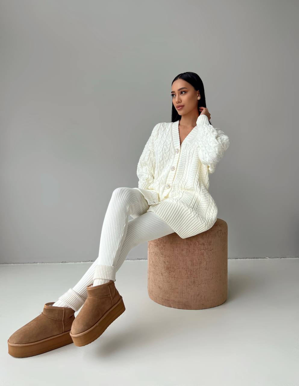 Woman sitting on a cylindrical stool wearing a white outfit and brown shoes against a plain background