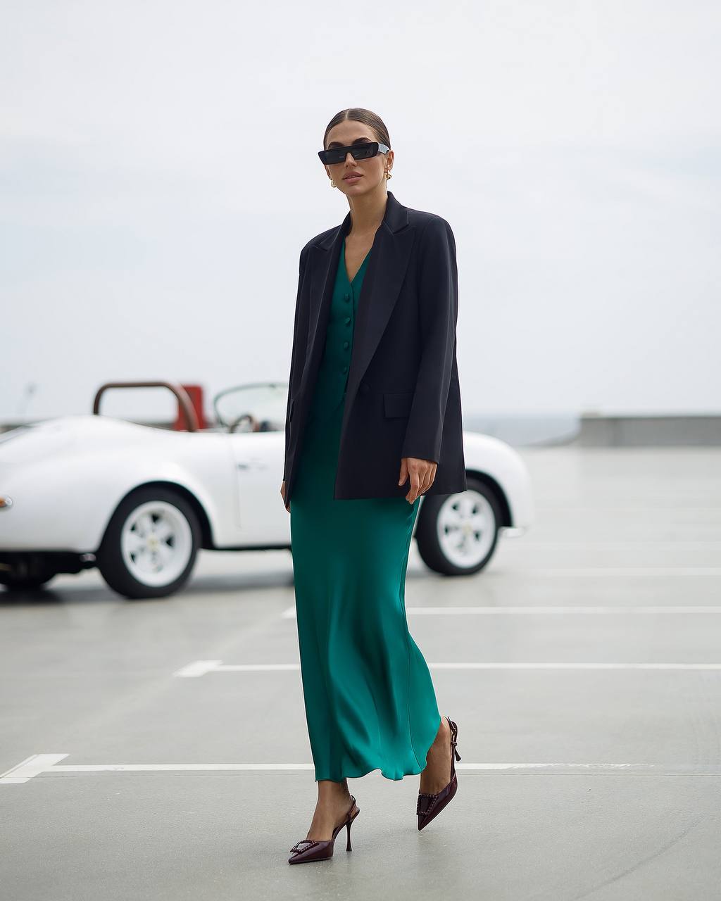 Woman in a green dress and black blazer standing in a parking lot with a white car in the background.