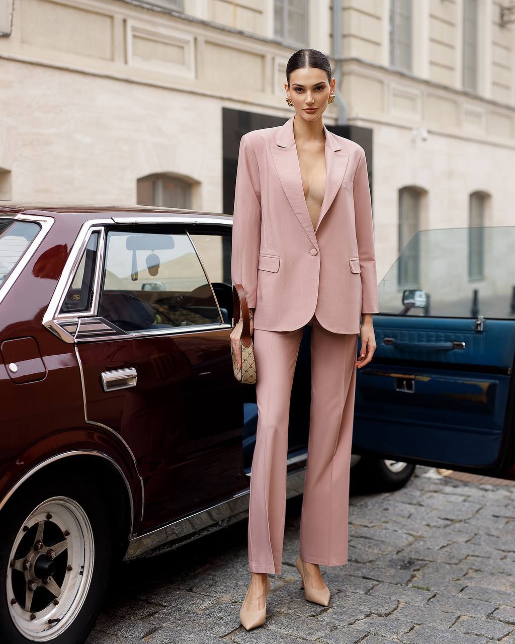 Woman in a pink suit standing next to a vintage car on a city street.