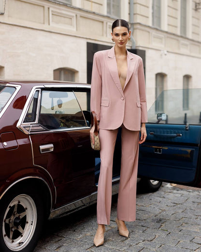 Woman in a pink suit standing next to a vintage car on a city street.
