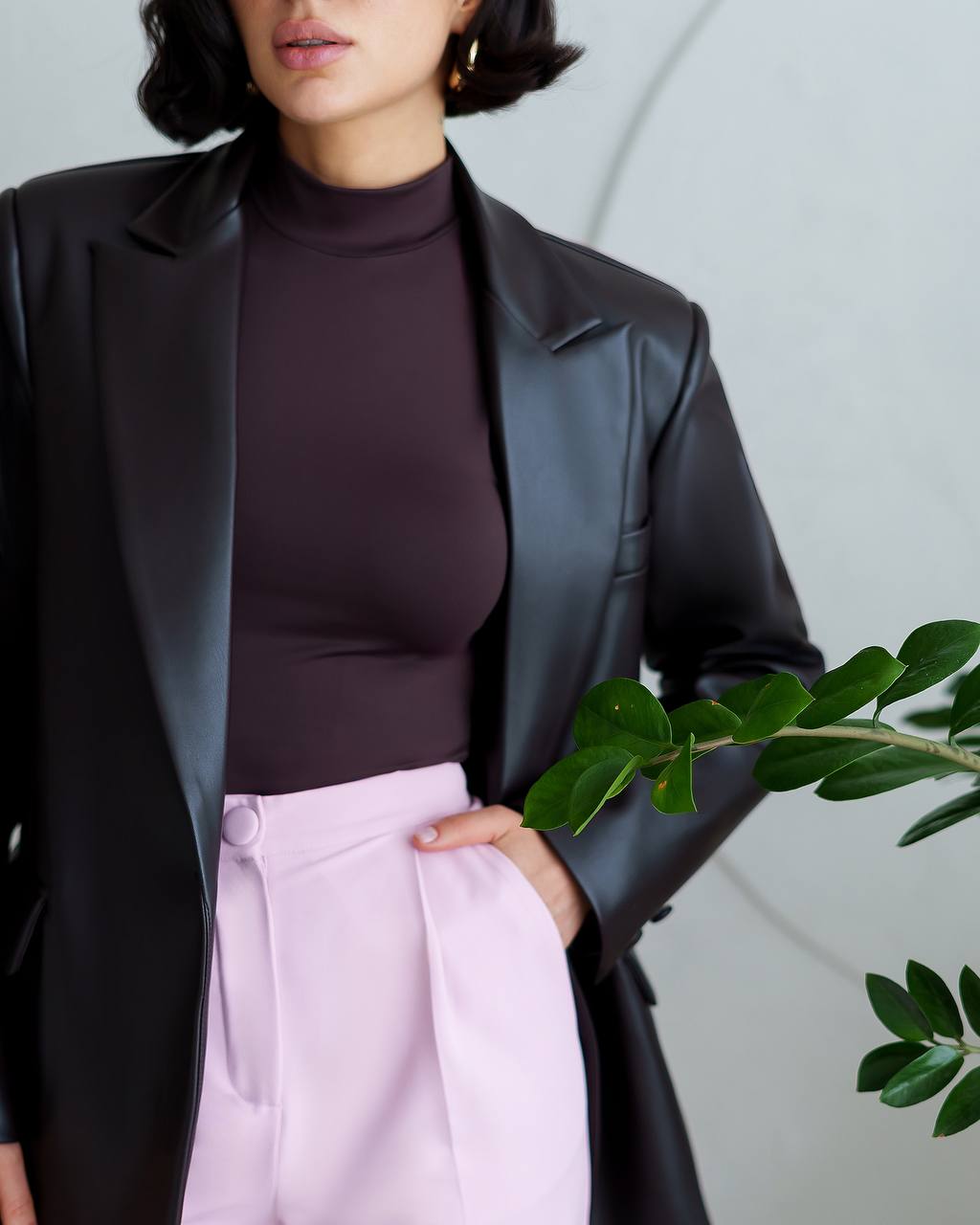 Person wearing a black leather jacket, dark turtleneck, and light pink pants with a plant in the foreground.