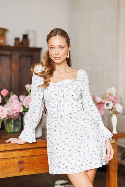 Woman wearing a white floral dress in a home setting with flowers and furniture.
