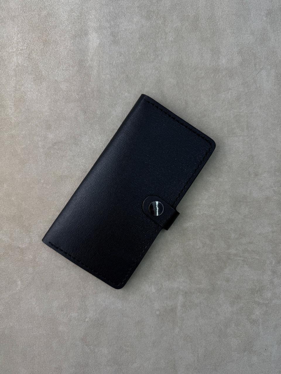 A black leather wallet with a snap-button closure, photographed from a top-down angle on a textured light gray surface.