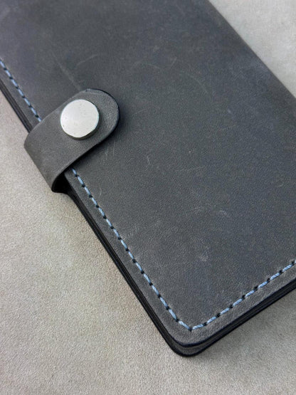 A close-up of a dark gray leather wallet or notebook cover with light blue stitching and a silver snap button closure, resting on a textured gray surface.