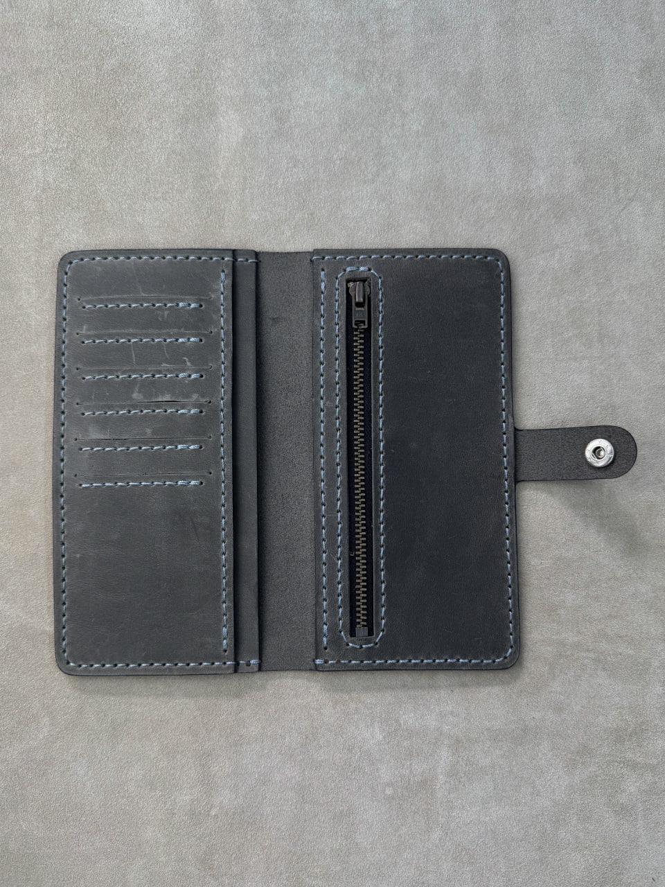 A dark gray leather wallet shown open, featuring multiple card slots on the left, a vertical zippered compartment in the center, and a strap with a silver snap button on the right, set against a textured gray background.