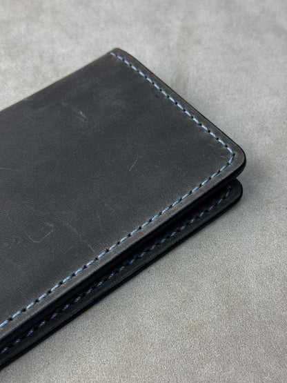 A close-up of a black leather wallet or card holder with light blue stitching, placed on a textured gray surface.