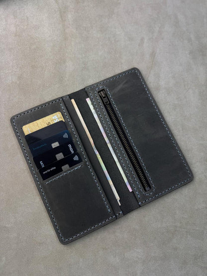 A dark gray leather wallet shown open on a textured gray surface, featuring card slots, a zippered pocket, and a pen loop.