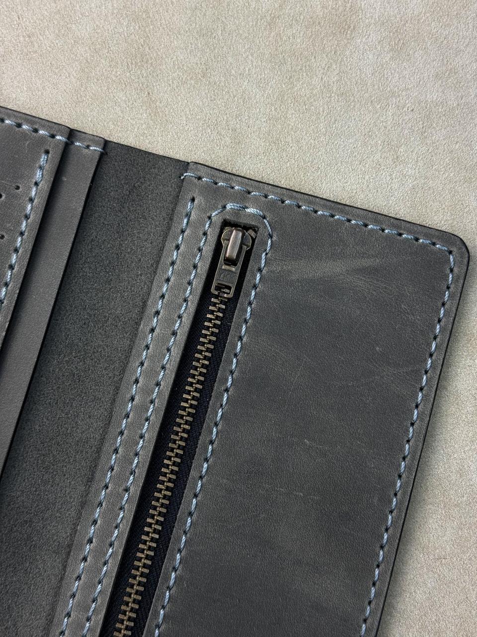Close-up of a dark gray leather wallet or folio case showing a zipper closure with light blue stitching on a neutral beige background.