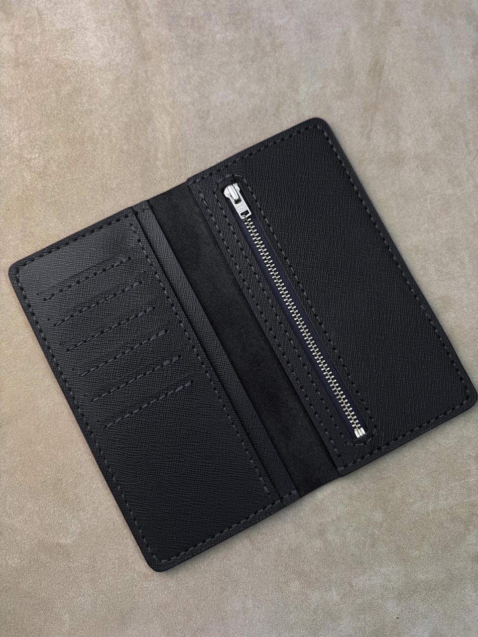 A black textured leather wallet shown open, displaying its interior layout with card slots, a central divider, and a zippered compartment, set against a neutral beige background.