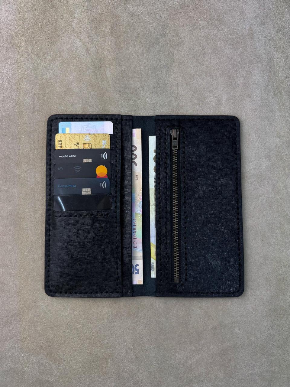 A black leather card holder wallet shown open, displaying card slots with sample cards, a zippered compartment, and cash, laid flat on a textured gray surface.