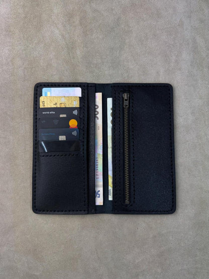 A black leather card holder wallet shown open, displaying card slots with sample cards, a zippered compartment, and cash, laid flat on a textured gray surface.