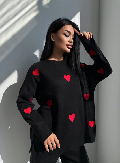 Woman wearing a black sweater with red heart patterns against a light gray wall.