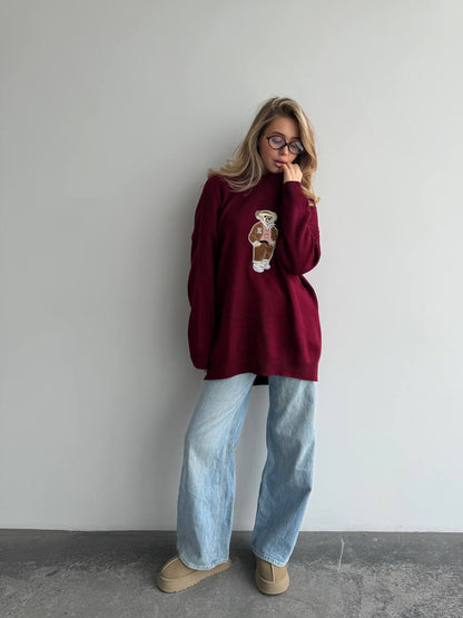 Person wearing a burgundy sweater with a graphic design, light blue jeans, and beige shoes against a plain background.