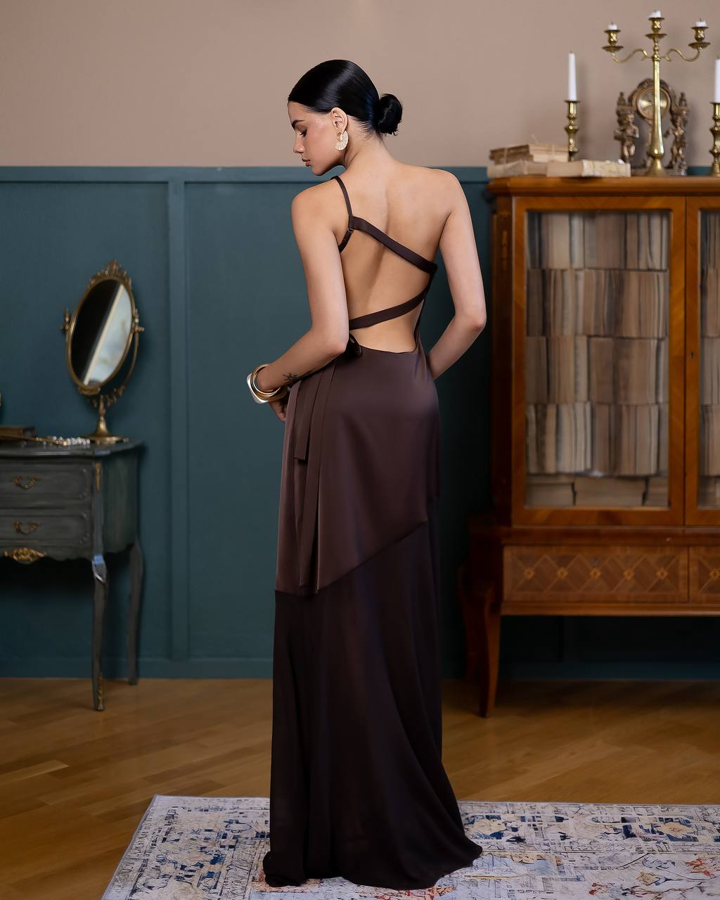 Woman in a long, dark brown dress standing in an elegant room with a mirror and wooden cabinet.