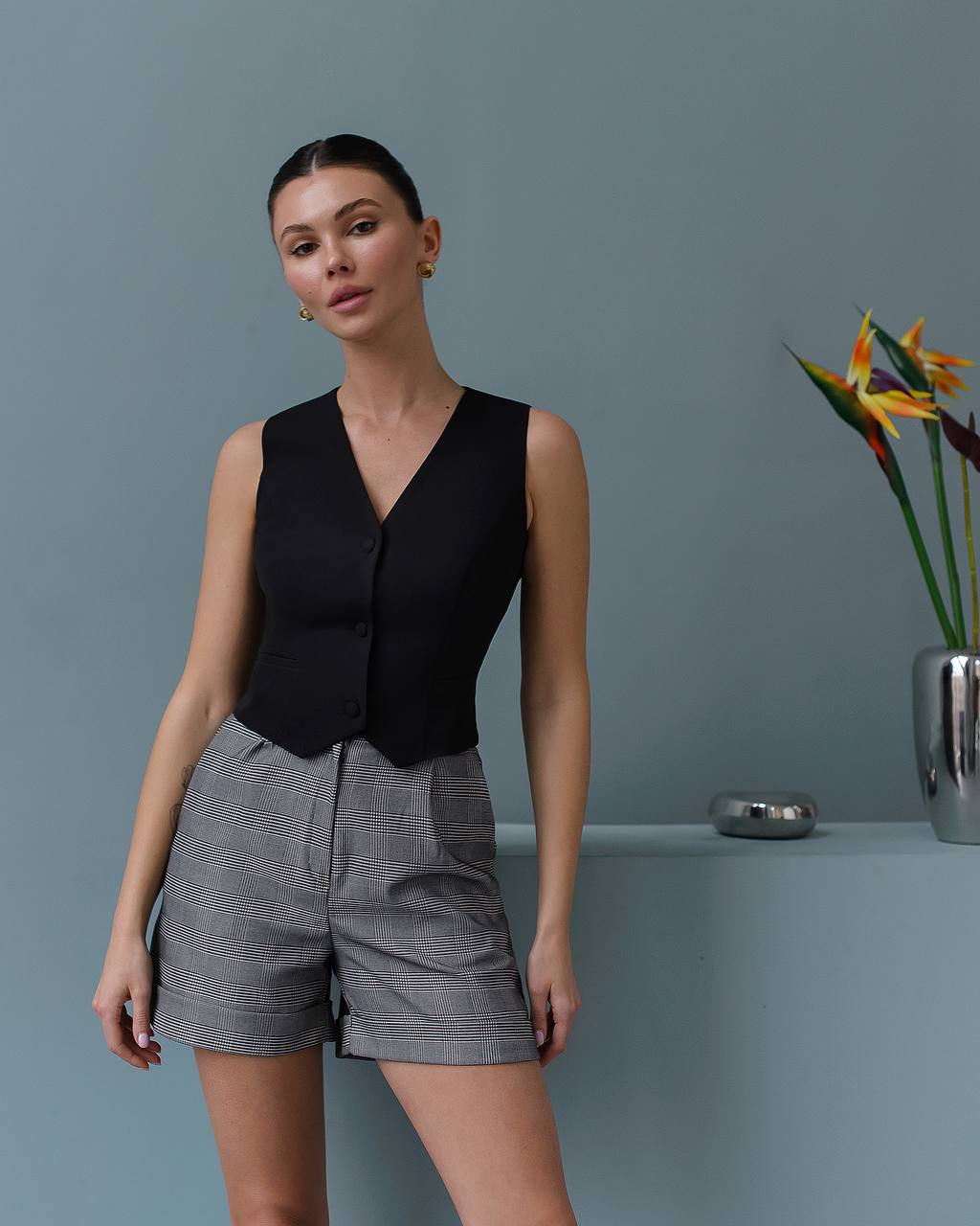 Woman wearing a black sleeveless top and gray shorts against a gray background with a plant.