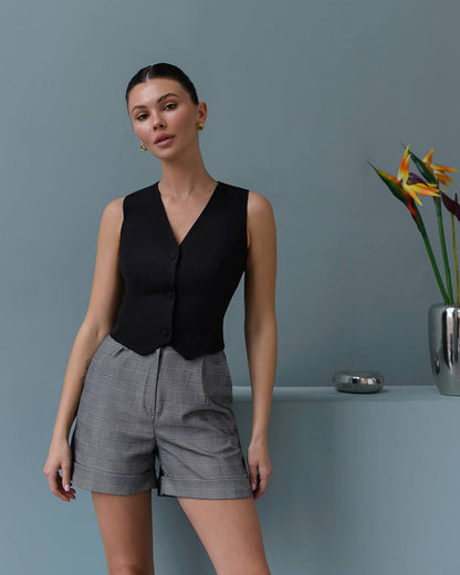 Woman wearing a black sleeveless top and gray shorts against a gray background with a plant.