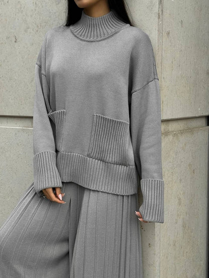 Person wearing a gray ribbed sweater and pants against a concrete wall.