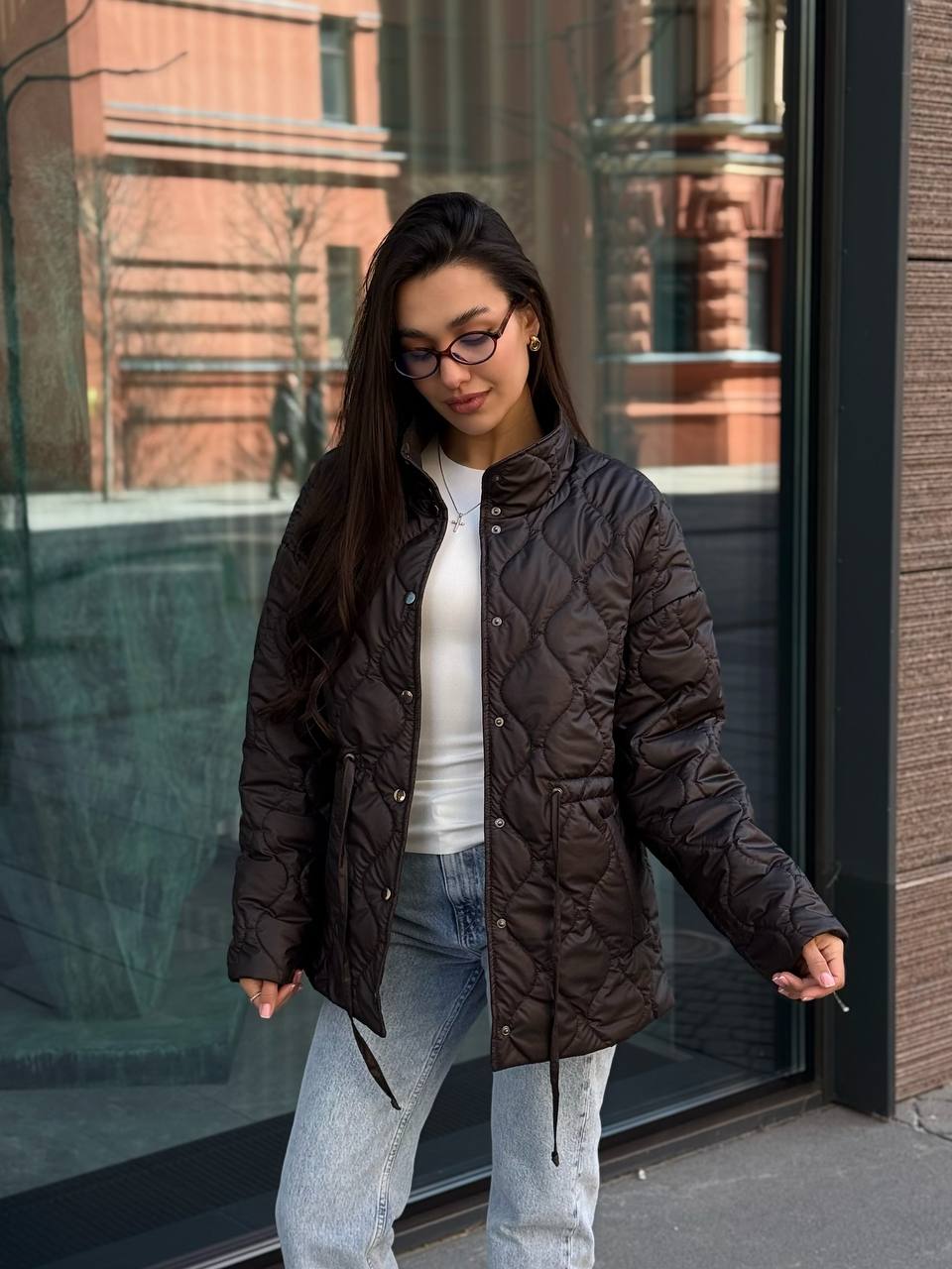 Woman wearing a dark quilted jacket and glasses standing in front of a building.