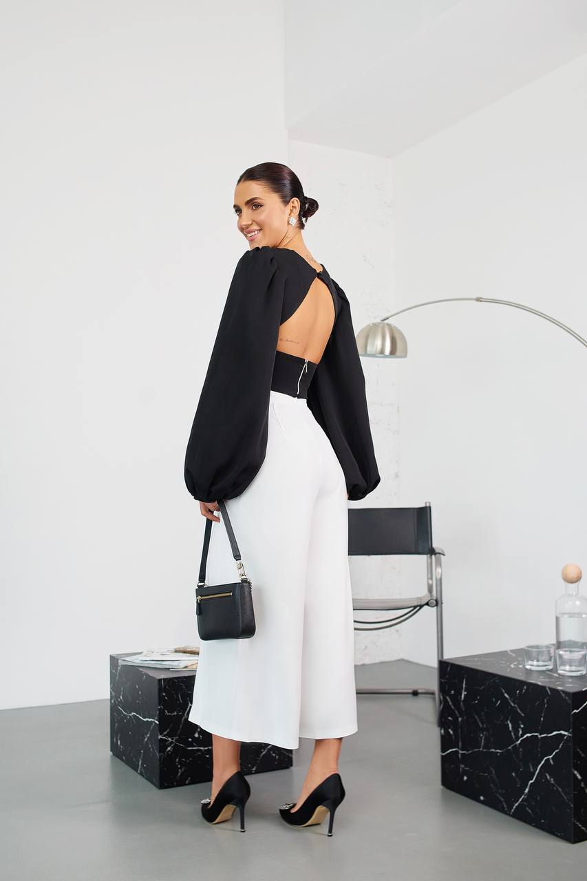 Woman in black top and white pants holding a black handbag in a minimalistic room.