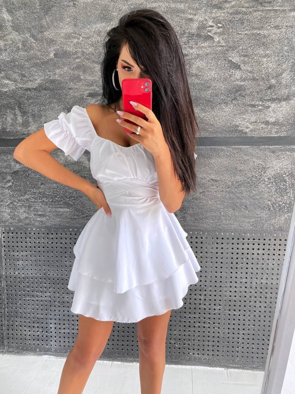 Woman in a white dress taking a mirror selfie against a gray wall.