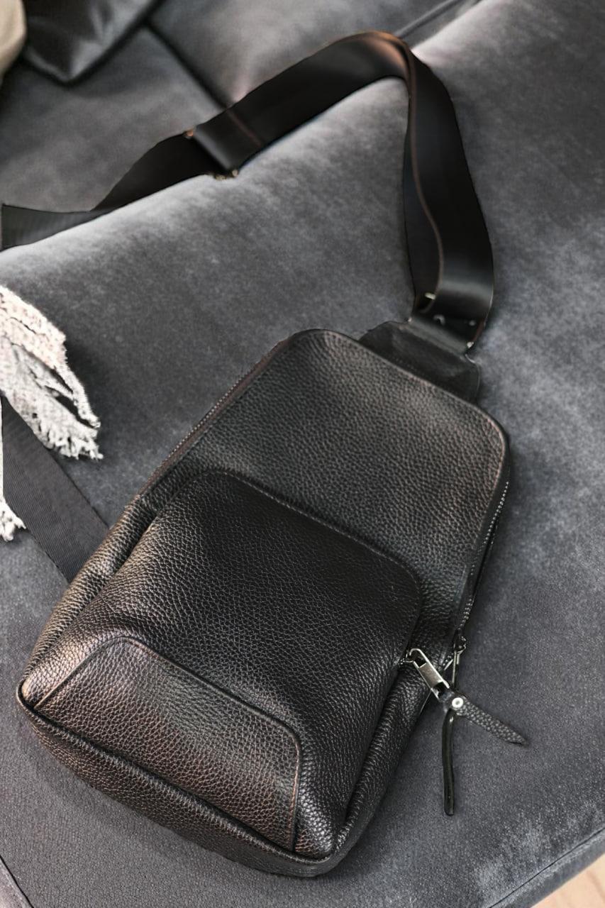 Black pebbled leather crossbody bag with a wide adjustable strap, resting on a gray velvet sofa.