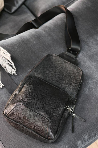 Black pebbled leather crossbody bag with a wide adjustable strap, resting on a gray velvet sofa.