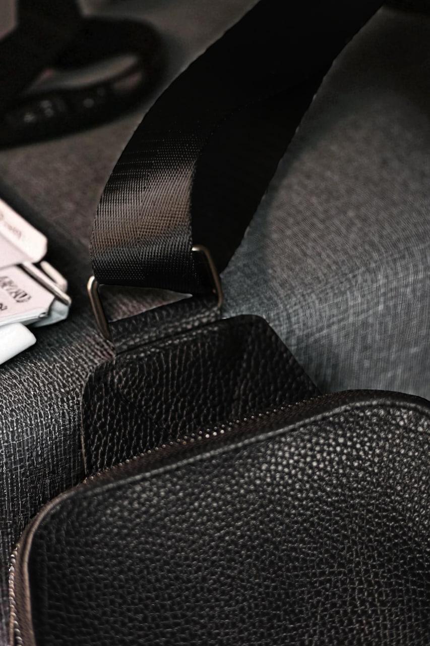 Close-up of a black pebbled leather crossbody bag strap attached to a dark gray textured bag, showing a metal buckle and fabric strap detail.