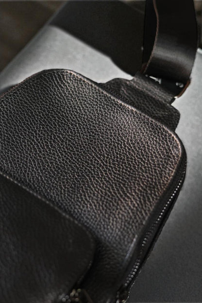 Close-up of a black textured leather bag showing the pebbled grain, zipper detail, and part of a dark strap against a dark gray background.