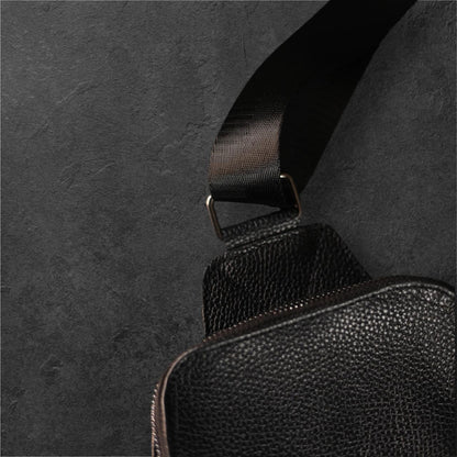 Close-up of a black pebbled leather bag with a matching black woven strap and metal hardware, set against a dark textured background.