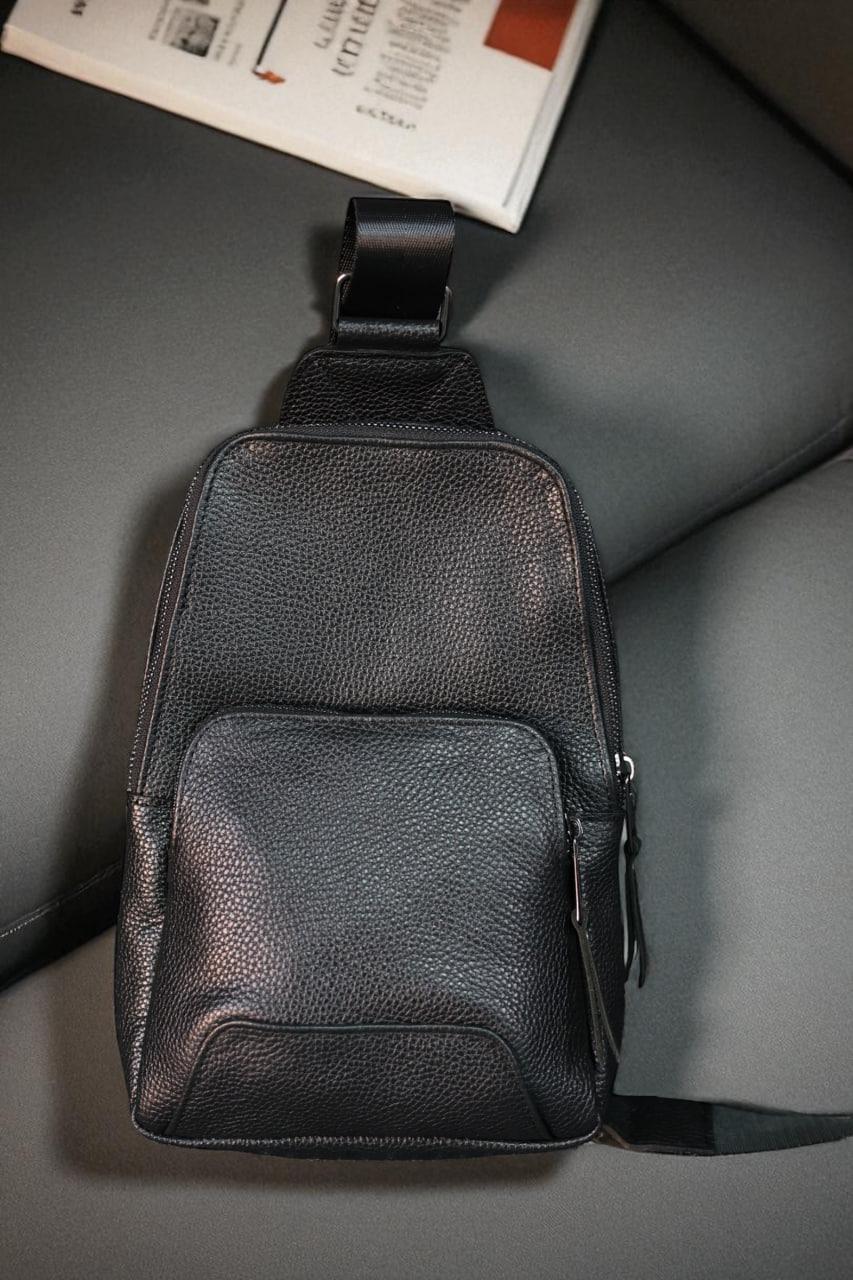 A black textured leather sling bag with a front zippered pocket and a top loop strap, resting on a dark gray leather seat next to a magazine.