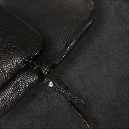 Close-up of a black pebbled leather bag with a zipper and leather pull tab on a dark textured background.