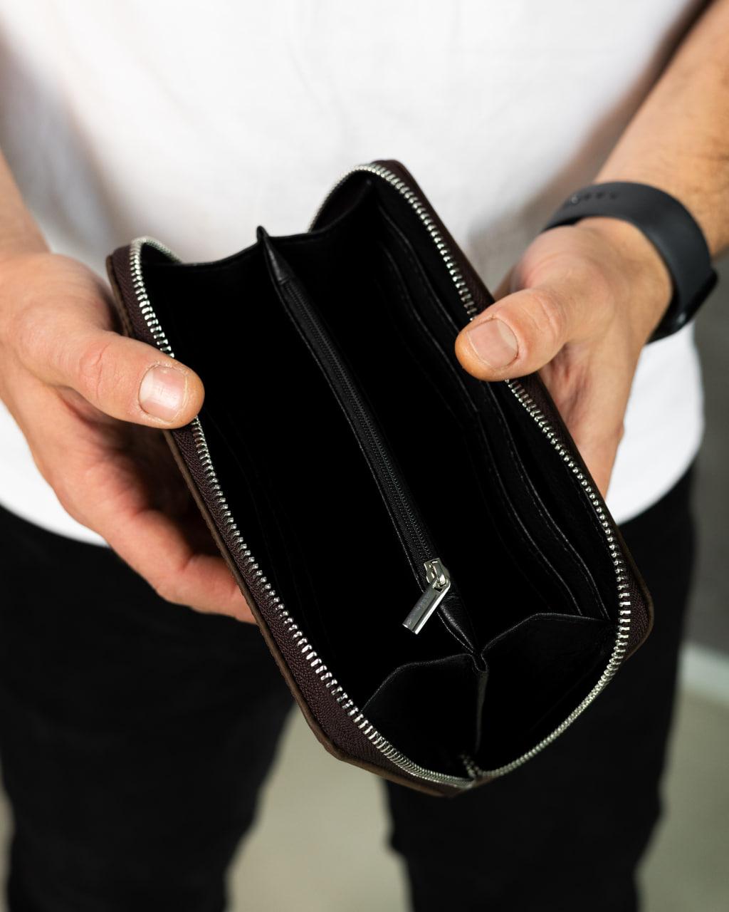 A person holding open a dark brown leather zip-around wallet or document case, showing the black interior compartments and silver zipper teeth.