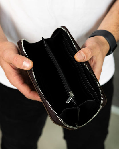 A person holding open a dark brown leather zip-around wallet or document case, showing the black interior compartments and silver zipper teeth.