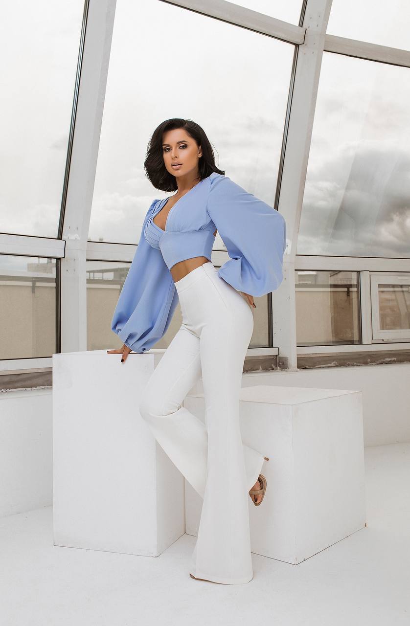 Woman in a light blue top and white pants posing in a modern indoor setting.