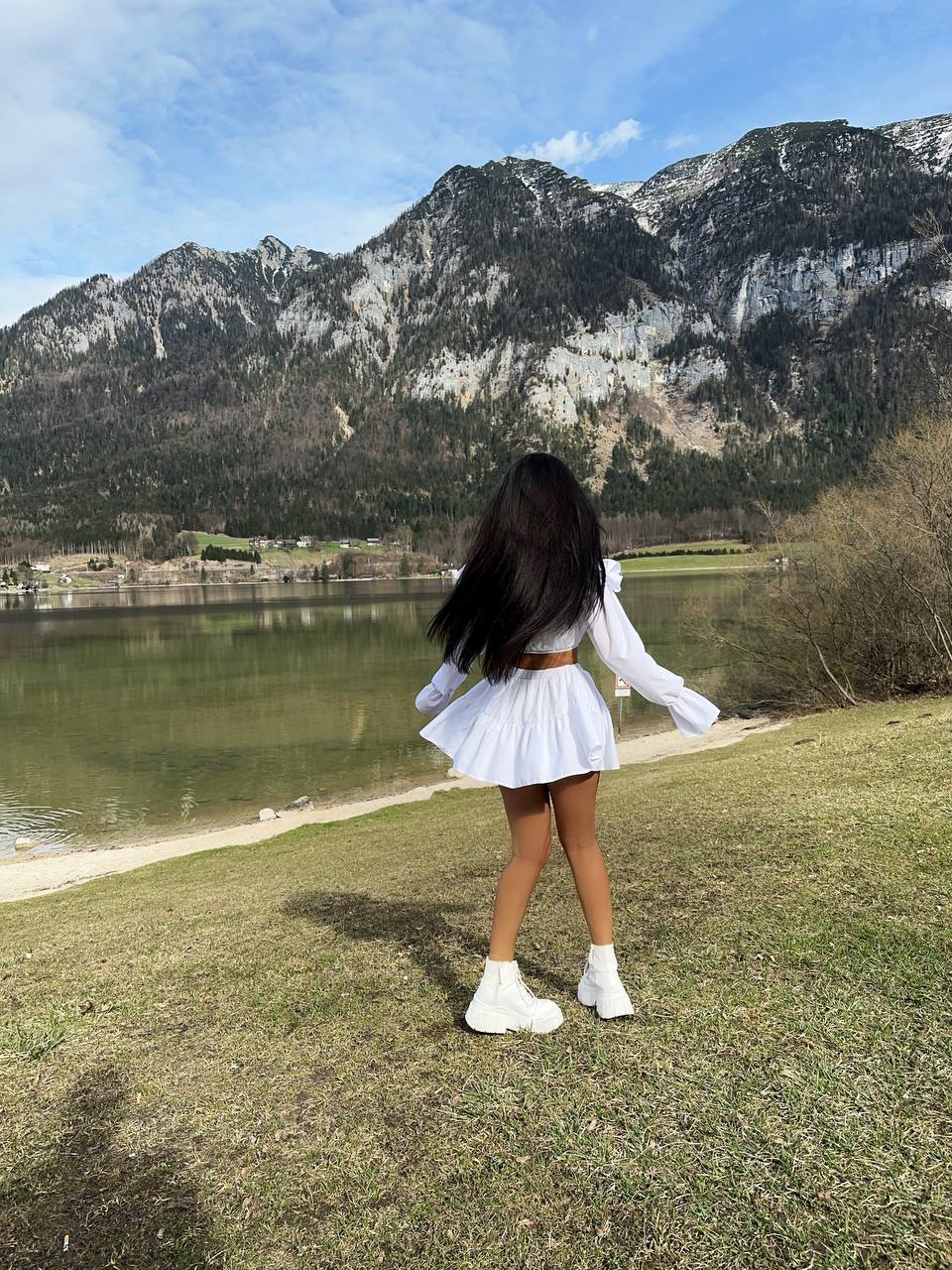 Person in a white outfit standing by a lake with mountains in the background