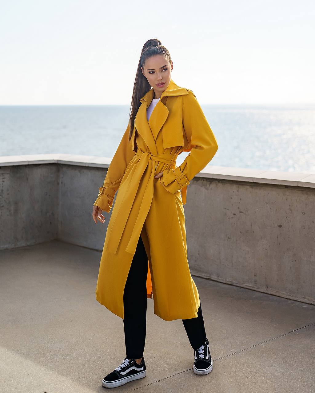 Woman wearing a yellow trench coat standing on a rooftop with ocean view