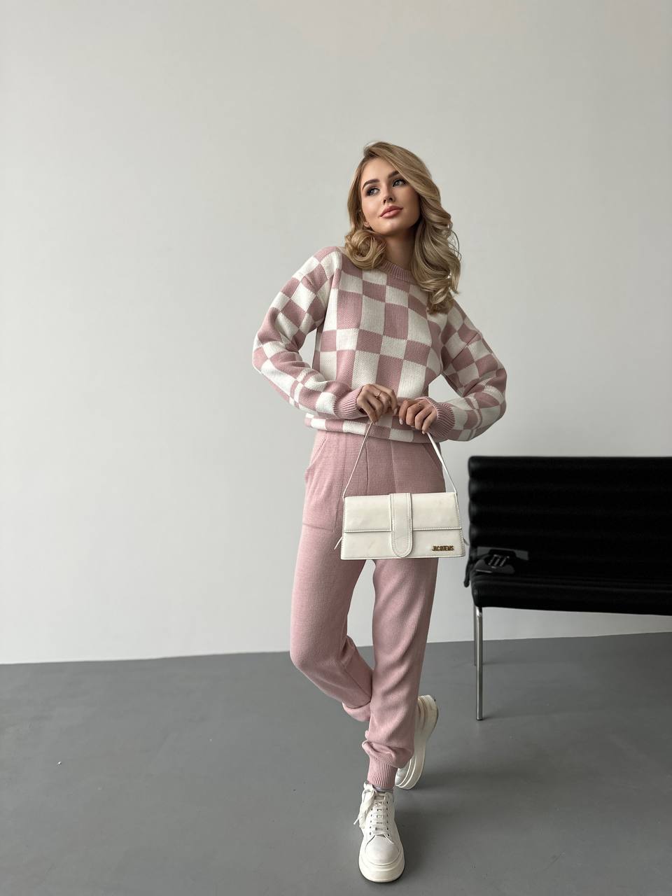 Woman wearing a pink and white checkered sweater and pants, holding a white handbag against a plain background.
