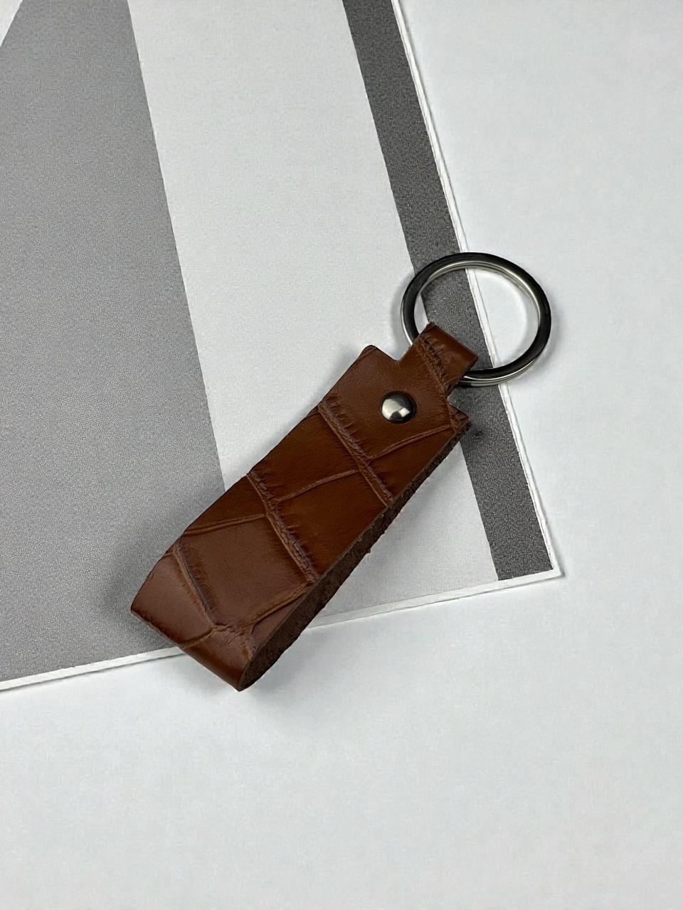 A brown crocodile-embossed leather keychain with a black metal split ring, resting on a modern gray and white geometric background.