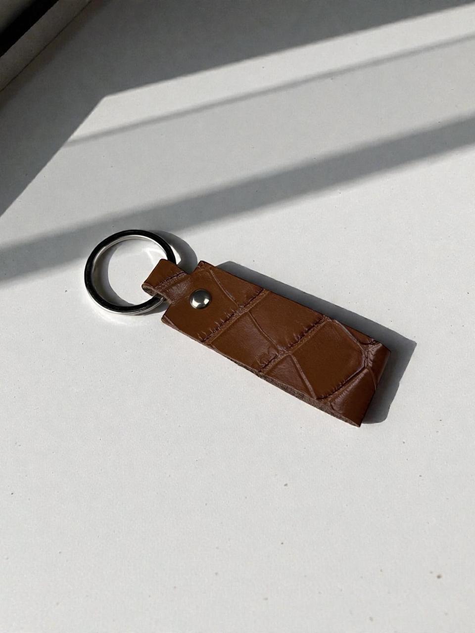 A brown leather keychain with a textured crocodile pattern, a silver snap button, and a black metal key ring, resting on a white surface with diagonal shadows.