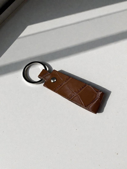 A brown leather keychain with a textured crocodile pattern, a silver snap button, and a black metal key ring, resting on a white surface with diagonal shadows.