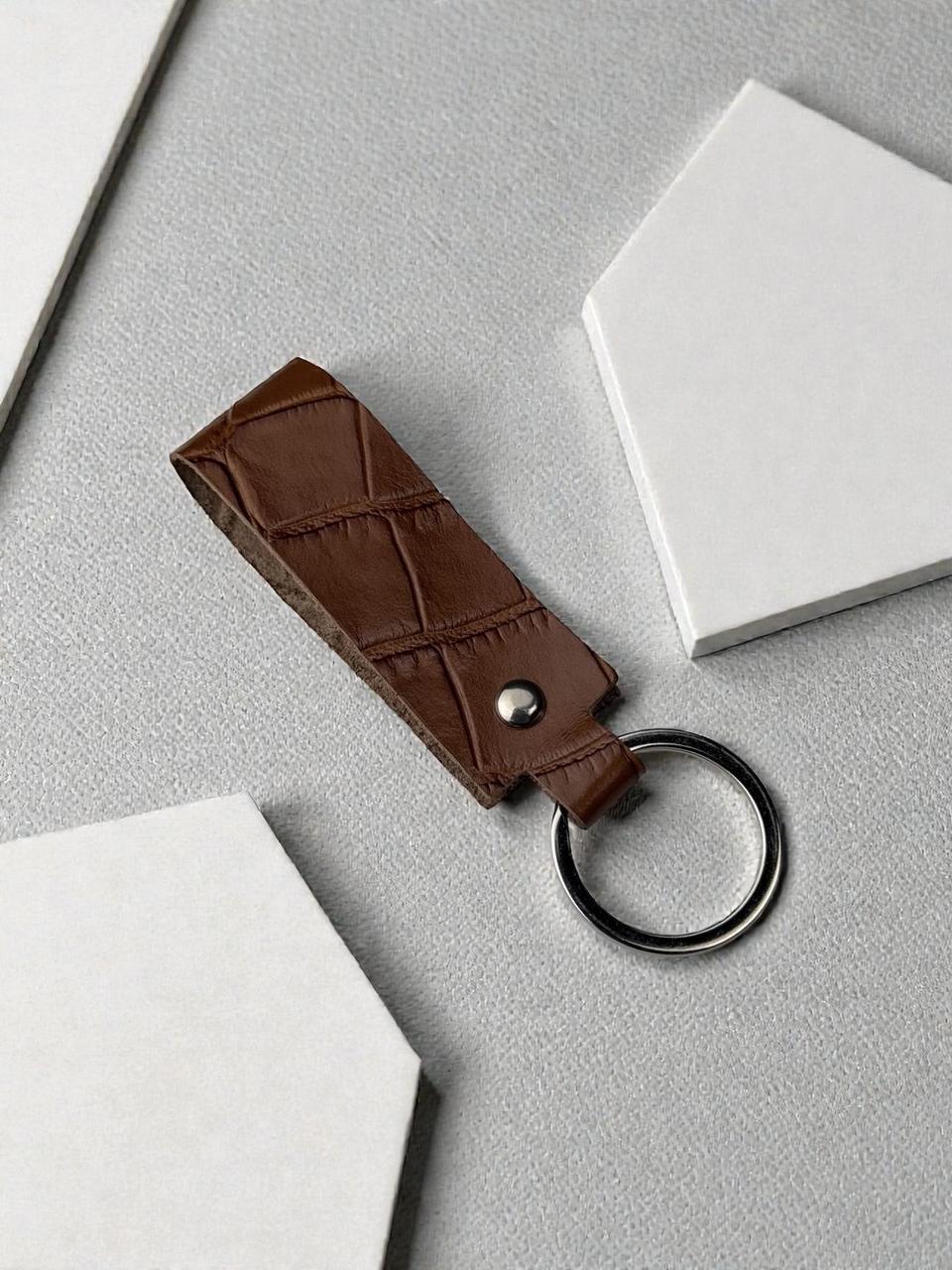 A brown crocodile-embossed leather keychain with a silver-tone metal rivet and a split key ring, displayed on a textured gray surface with white geometric props.