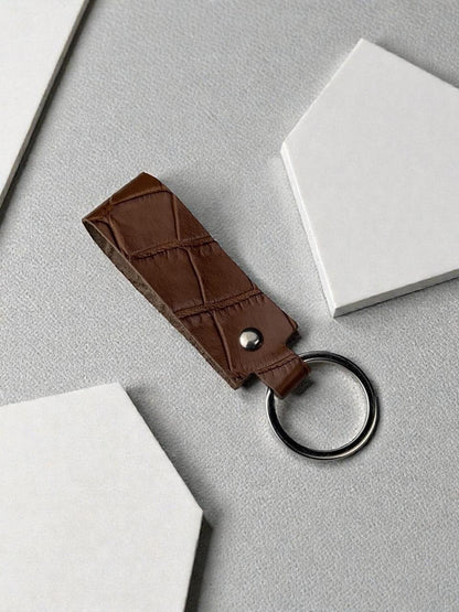 A brown crocodile-embossed leather keychain with a silver-tone metal rivet and a split key ring, displayed on a textured gray surface with white geometric props.
