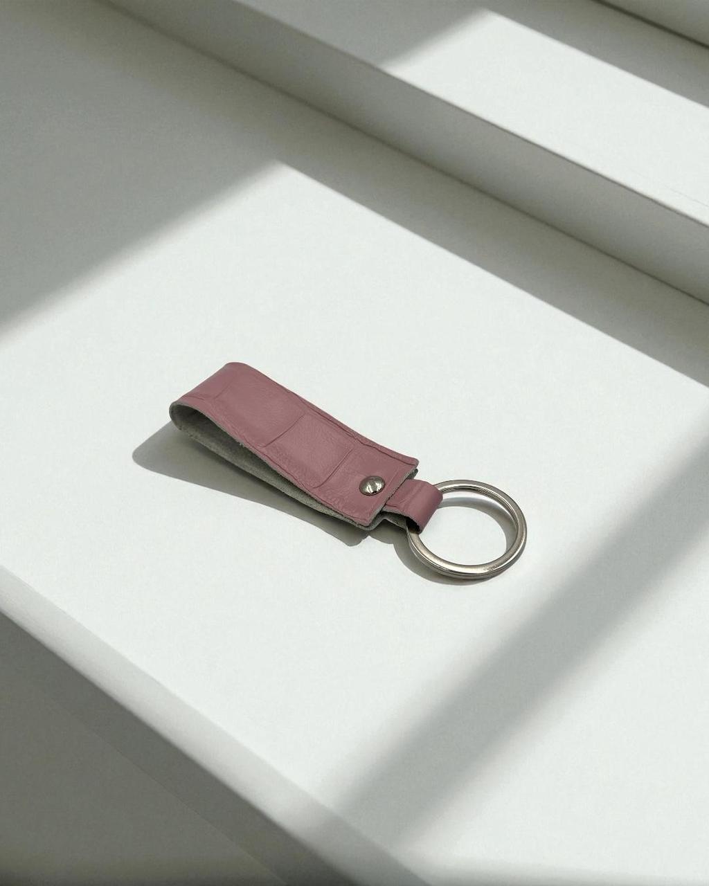 A dusty rose leather keychain with a silver metal key ring, resting on a white surface with soft diagonal shadows.