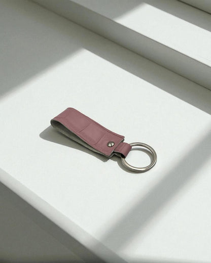 A dusty rose leather keychain with a silver metal key ring, resting on a white surface with soft diagonal shadows.