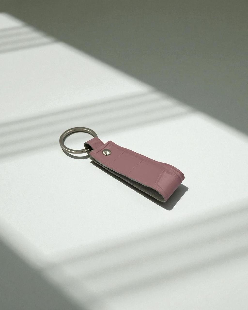 A dusty pink leather keychain with a silver metal split ring, resting on a white surface with soft diagonal shadows.