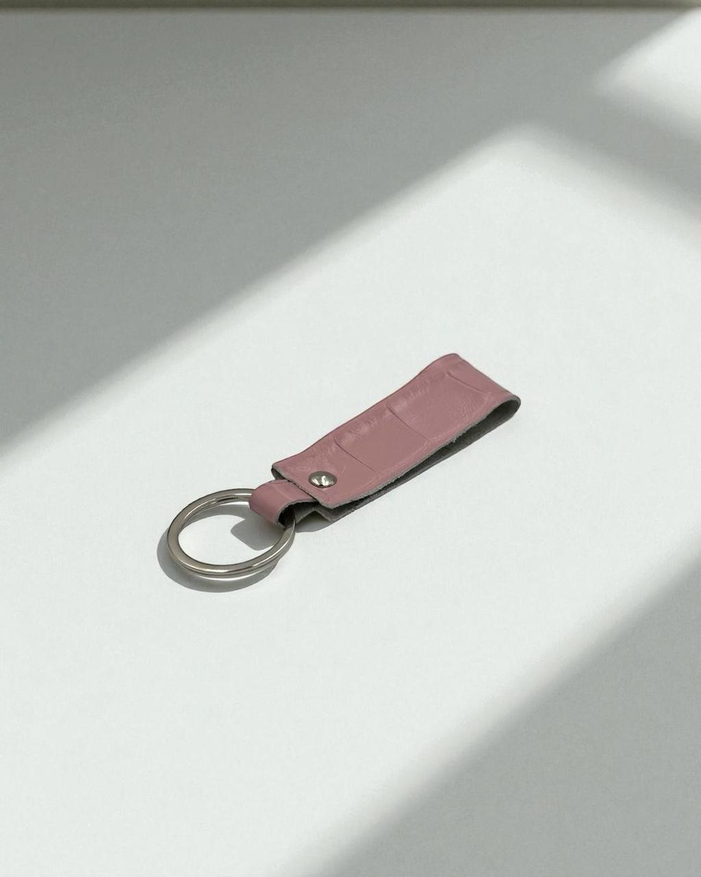 A pink faux-croc leather keychain with a silver metal split ring, resting on a white surface with soft diagonal shadows.