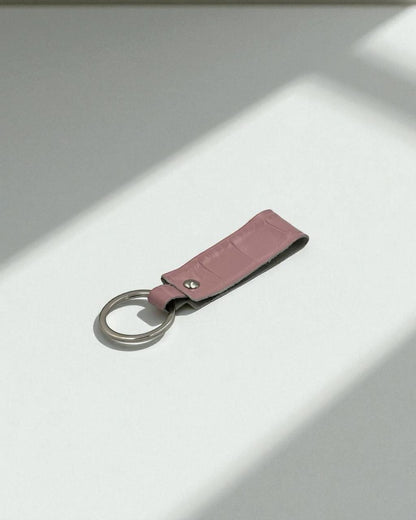 A pink faux-croc leather keychain with a silver metal split ring, resting on a white surface with soft diagonal shadows.