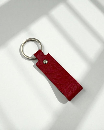 A red textured leather keychain with a silver metal key ring and rivet, photographed on a white surface with soft window shadow patterns.
